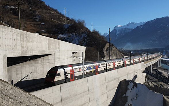 Ein Doppelstockzug fährt im gebirgigen Gelände auf einem Viadukt in einen Tunnel ein.